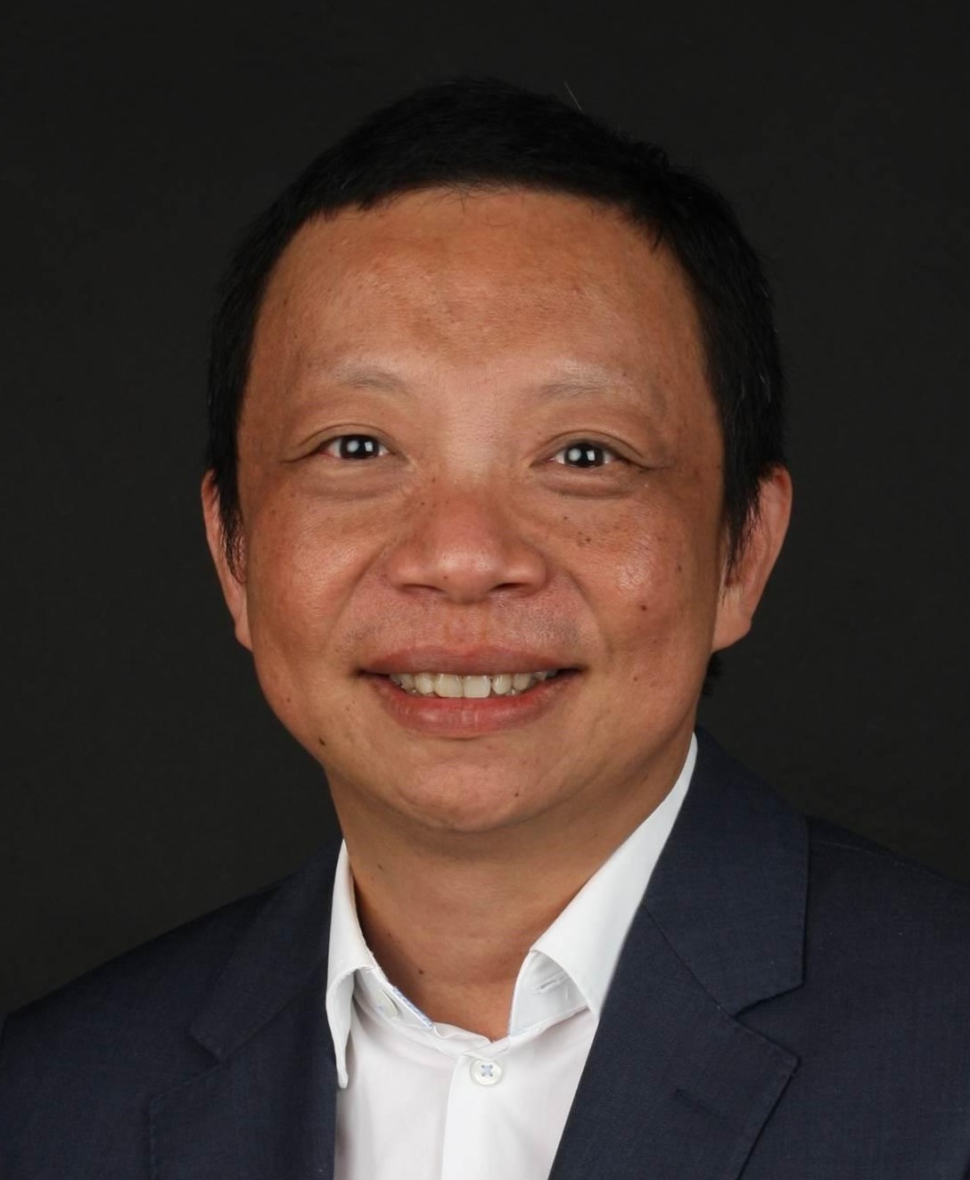 A person wearing a suit and white shirt is smiling against a dark background.